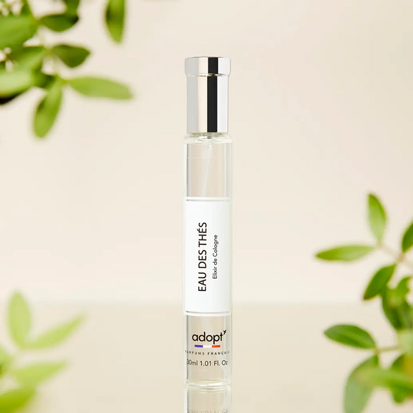 Adopt Eau Des Thés Elixir de Cologne 30ml bottle with a silver cap, surrounded by green leaves in a soft-focus background