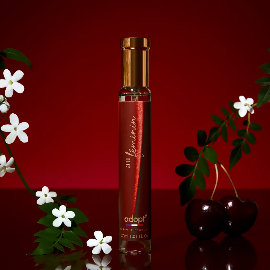 Adopt Au Feminin perfume bottle with a red label and gold cap, surrounded by white jasmine flowers and cherries