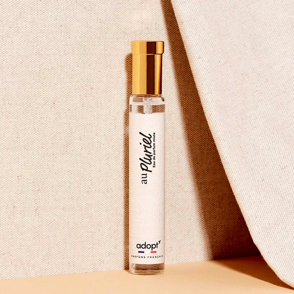 Adopt Au Pluriel Eau de Parfum Mixte in a clear glass bottle with a gold cap, standing upright on a textured background