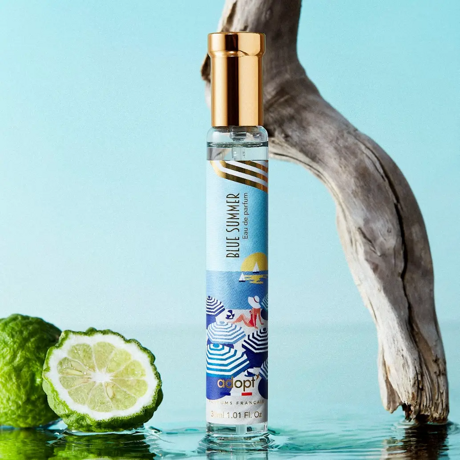 Adopt Blue Summer eau de parfum with a gold cap and a beach-themed label, standing in water with a sliced lime and driftwood