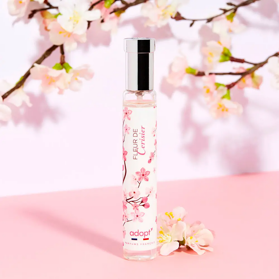 Adopt Fleur De Cerisier perfume bottle with cherry blossom design, displayed against a pink background with floral branches