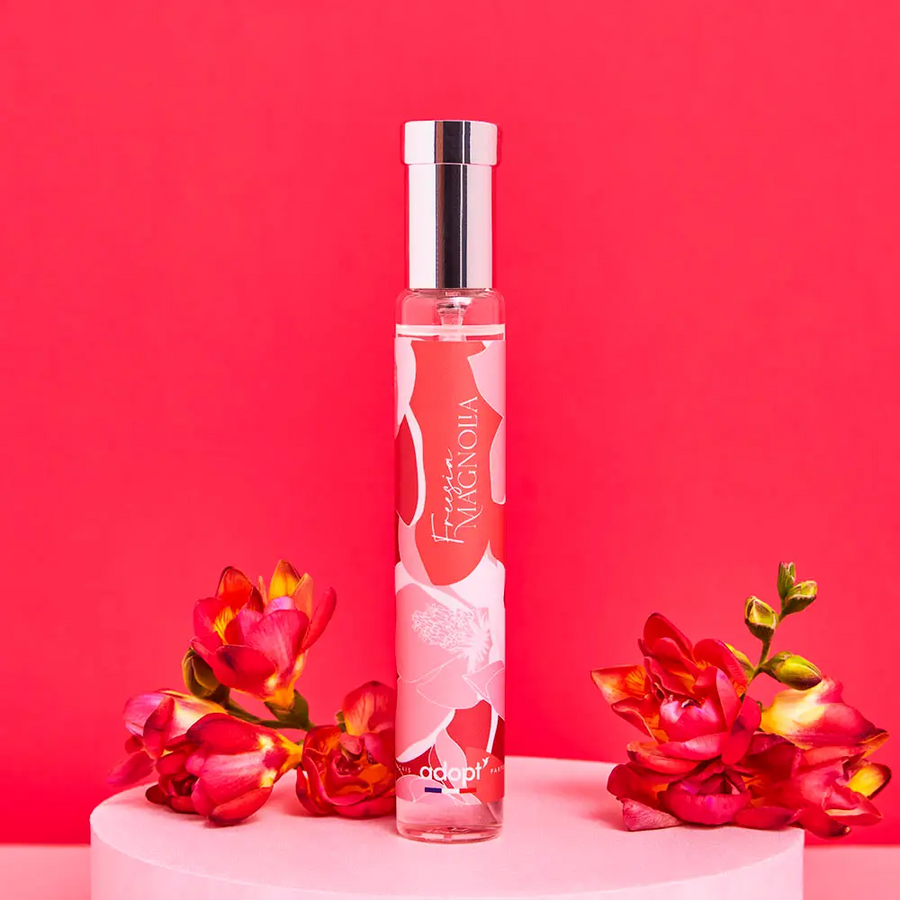 Adopt Freesia Magnolia perfume bottle with a floral design, standing on a pink pedestal with red flowers on a red background