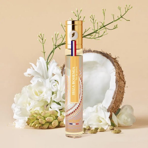 Adopt Ibiza Bohemia Eau de Parfum bottle with a gold cap, surrounded by white flowers, green leaves, and a coconut half