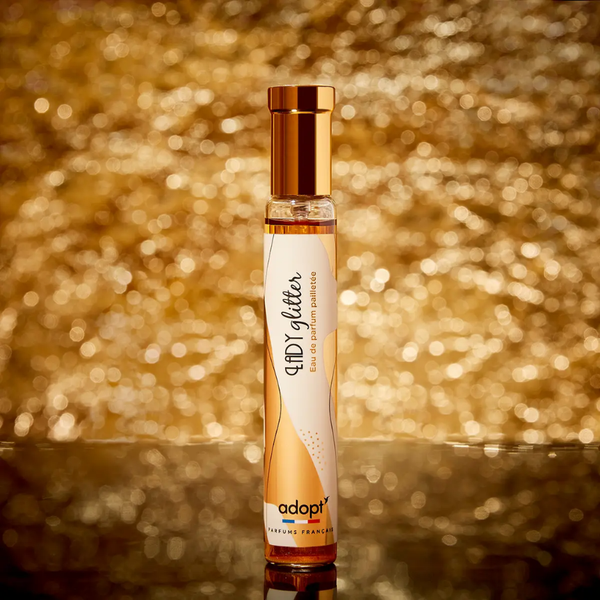Adopt Lady Glitter Eau de Parfum pailletée in a clear bottle with a gold cap, set against a shimmering gold background