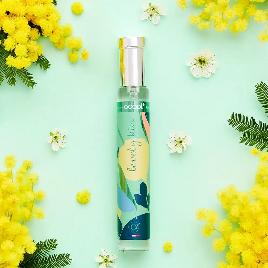 Adopt Lovely Kiss perfume bottle with a colorful label, surrounded by yellow mimosa flowers and white blossoms on a light