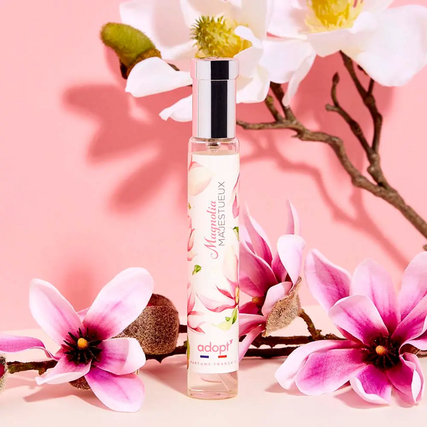 Adopt Magnolia Majestueux perfume bottle with a silver cap, surrounded by pink and white magnolia flowers on a pink