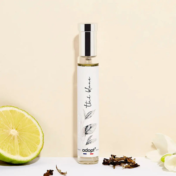 Adopt The Blanc (White Tea) perfume bottle with a silver cap and white label, next to a lime and dried tea leaves